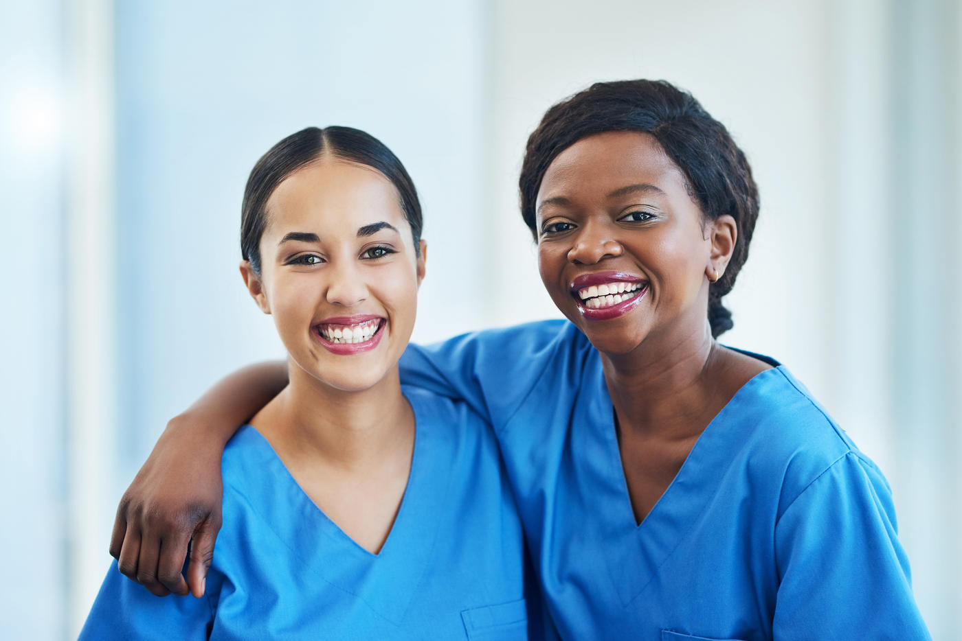 Two smiling women in scrubs, one with her arm around the other